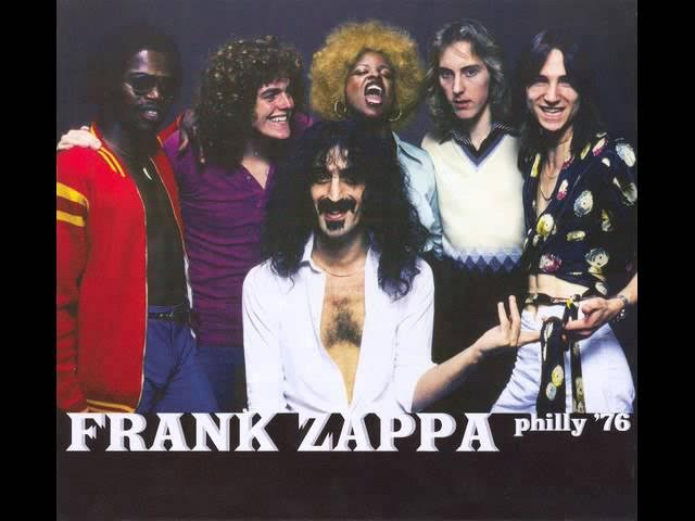 Frank Zappa ve grubu, 1976'da bir etkinlikte poz verirken, arka planda altı kişiyle birlikte gülümsemektedir.