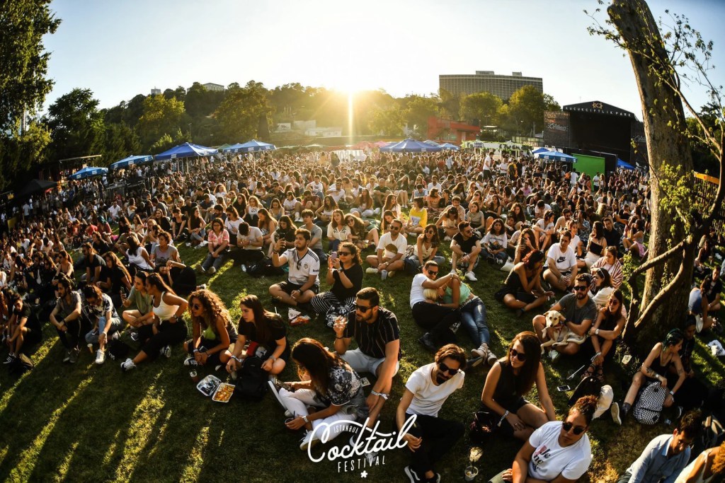 İstanbul Cocktail Festival 2025'te, Lifepark'ta kalabalık bir seyirci topluluğu oturarak konser izliyor. Güneşli bir gün, birçok kişi şort ve tişört giymiş, bazıları içecekler tutuyor.