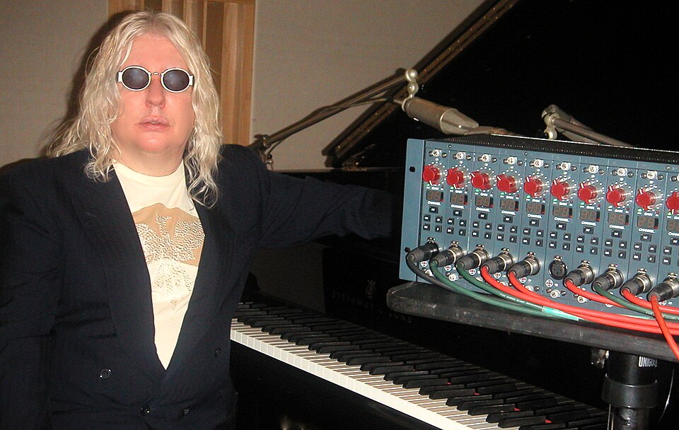 Roy Thomas Baker, müzik prodüktörü, stüdyoda mikrofon ve piyanonun önünde poz veriyor, gözlük takıyor.