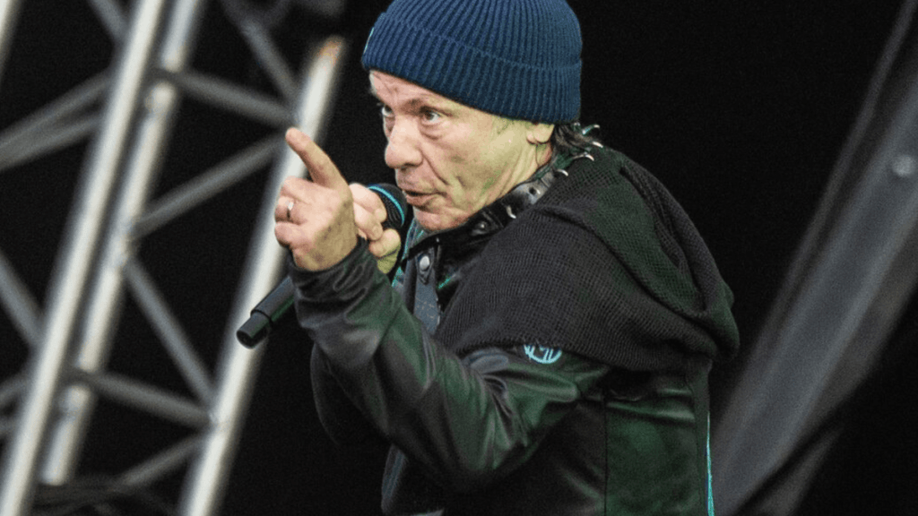 Bruce Dickinson, sahnede performans sergileyerek seyirciyle etkileşimde bulunan bir müzisyen, elini işaret ederek konuşuyor.