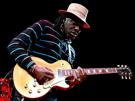 Joe Louis Walker, sahnede elektrikli gitar çalarken, şapka takıyor ve çizgili bir kazak giymektedir.