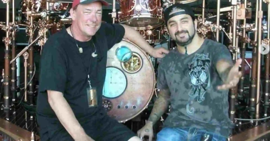 Mike Portnoy ve bir diğer kişi, büyük bir davul setinin önünde gülümseyerek oturuyor.